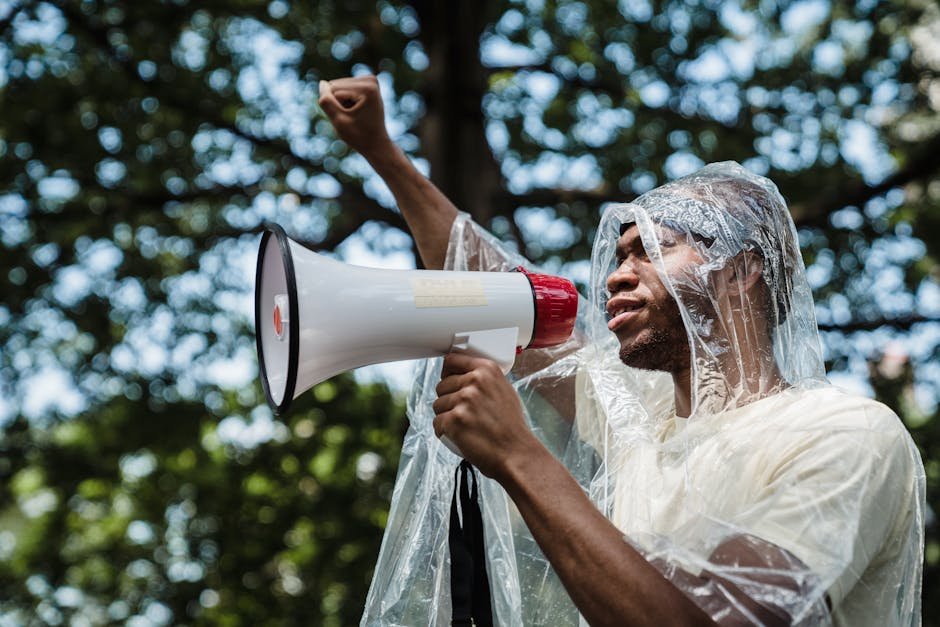 pexels-photo-8899045-8899045 A man wearing a raincoat raises a fist and uses a megaphone to protest outdoors.