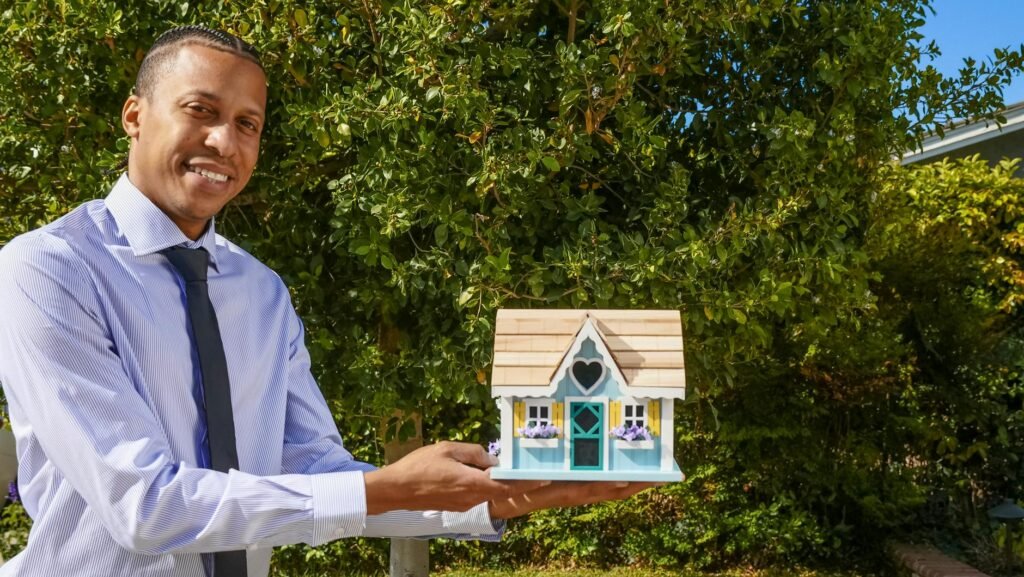Smiling real estate agent presenting a miniature house model outdoors against a background of greenery.